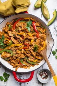 A serving dish of vegan chicken fajitas with a wooden spoon on a white wooden table.