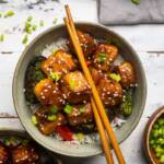 A bowl of Vegan General Tso's Tofu over rice with broccoli and chopsticks on top.