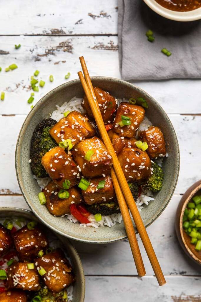 A bowl of Vegan General Tso\'s Tofu over rice with broccoli and chopsticks on top. 