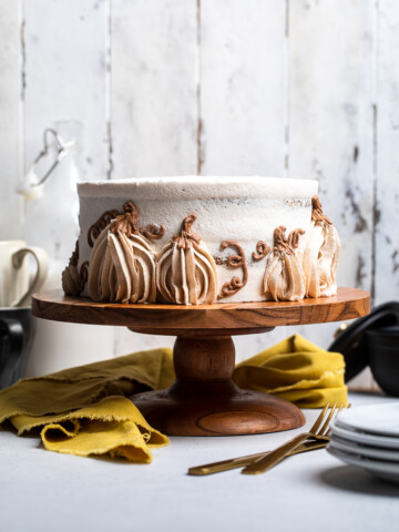 An uncut layered vegan pumpkin cake on a cake stand with pumpkins designed with frosting.