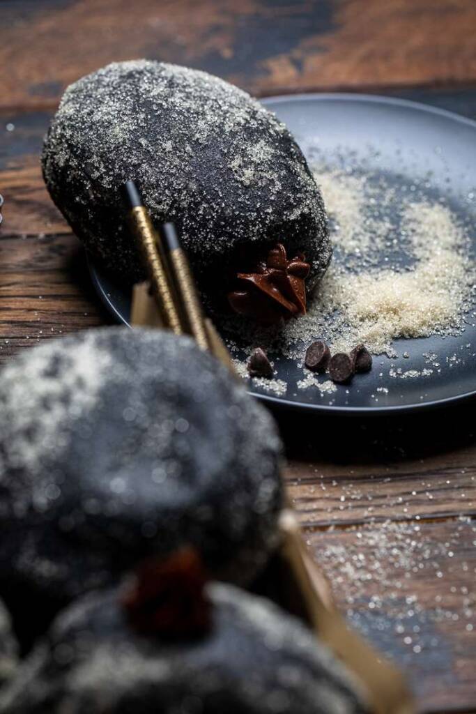 A black plate of sugar with a black chocolate vegan brioche donut on the side.
