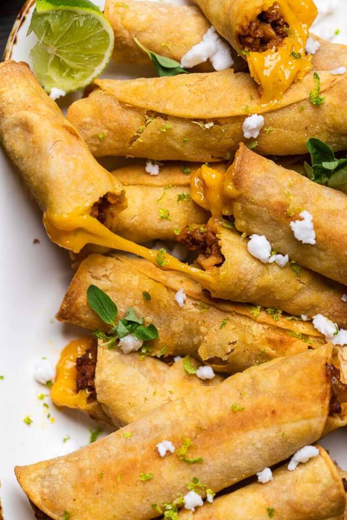 A few cheesy baked vegan taquitos on a plate with crumbled cheese and herbs.