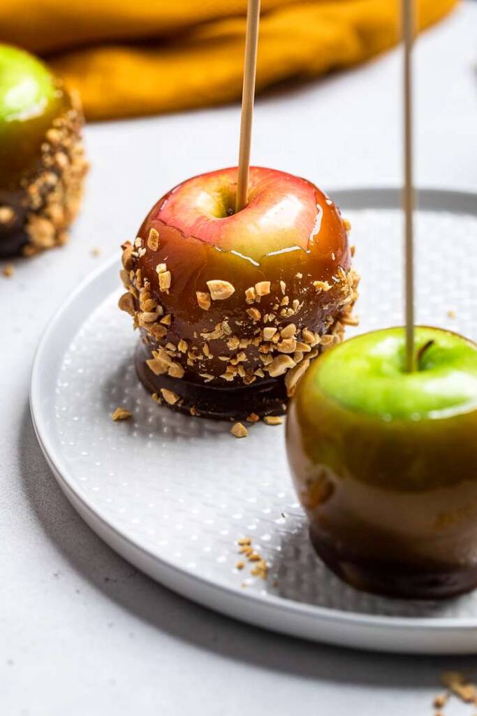 A white platter of one green vegan caramel apple and one red with chopped peanuts.