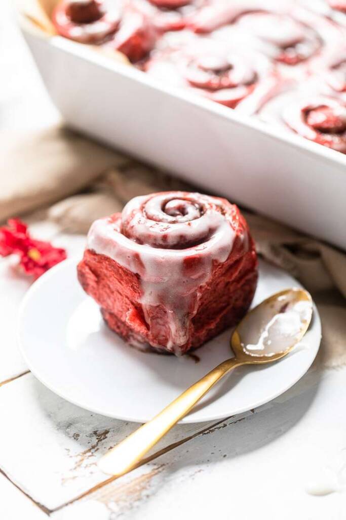 A single Vegan red velvet cinnamon roll on a white plate with a gold spoon.