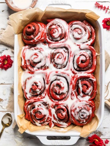 A white pan of vegan red velvet cinnamon rolls with melted cream cheese frosting.