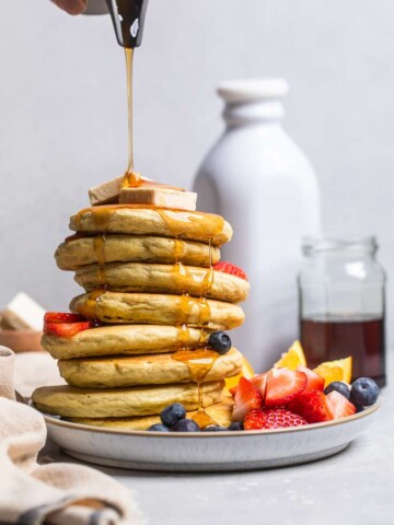 A stack of fluffy vegan protein pancakes with syrup drizzling from top on a white plate.