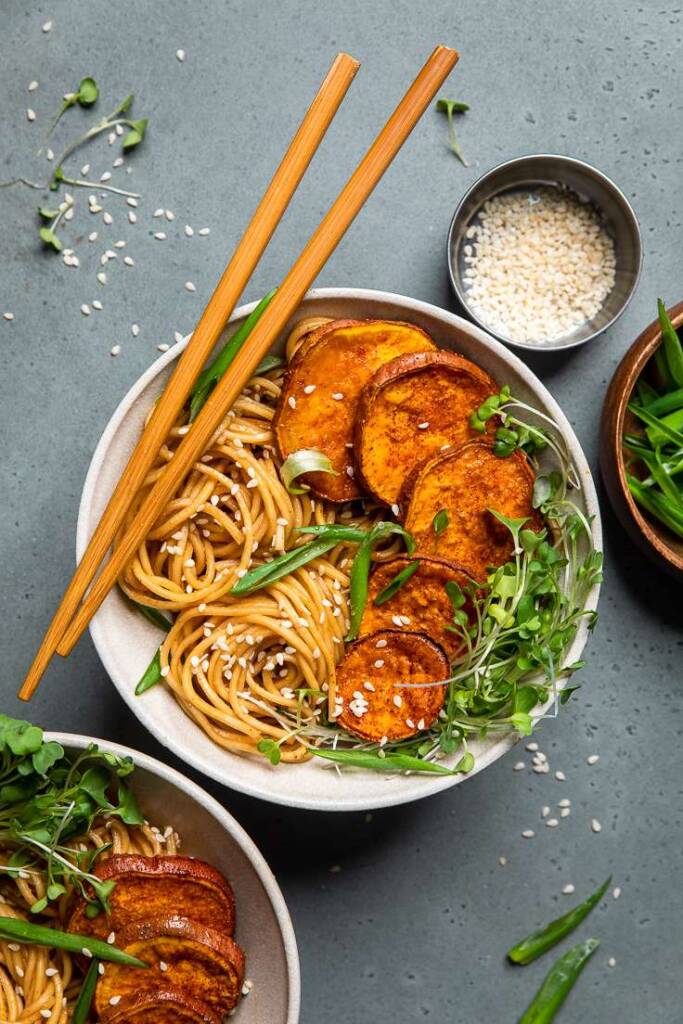 a white bowl of easy vegan sesame garlic noodles, sliced sweet potatoes, and microgeens.
