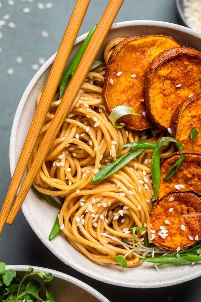 Vegan sesame garlic noodles in a white bowl with slices sweet potatoes and chopsticks. 