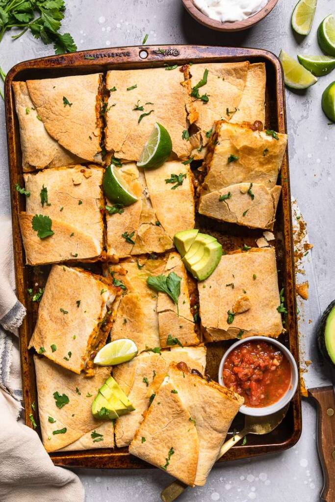 Cut Vegan sheet pan quesadillas with a bowl of salsa, lime wedges and sliced avocados. 