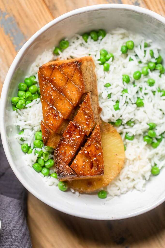 A piece Vegan brown sugar glazed tofu overtop rice and peas with a slice of pineapple. 