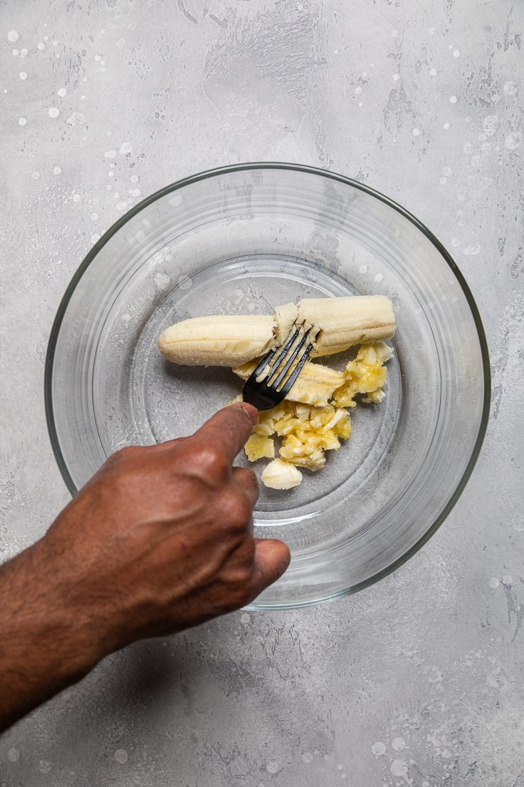 Hand mashing banana with a fork in a clear bowl.