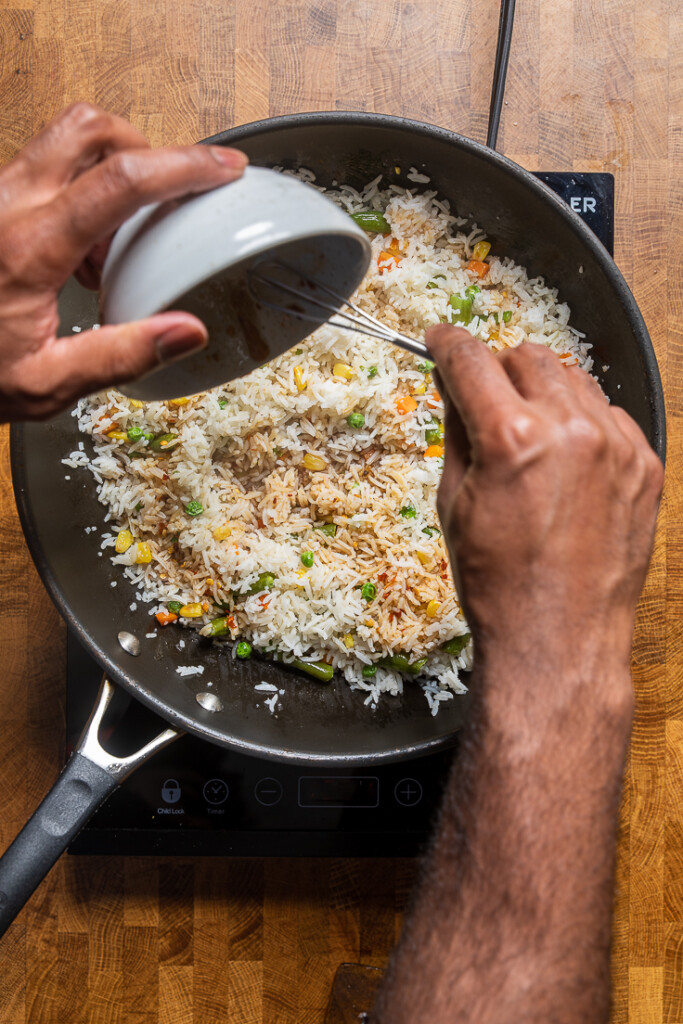 Hands pouring soy sauce mixture into skillet of rice and vegetables. 