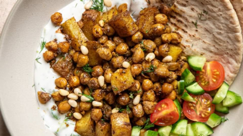a bowl filled with tzatziki, roasted spiced chickpeas and potatoes, tomatoes, cucumbers, topped with pine nuts and quartered pitas on the side.