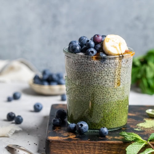 matcha chia pudding in a glass jar topped with fresh blueberries and banana slices with maple syrup drizzling down the side.