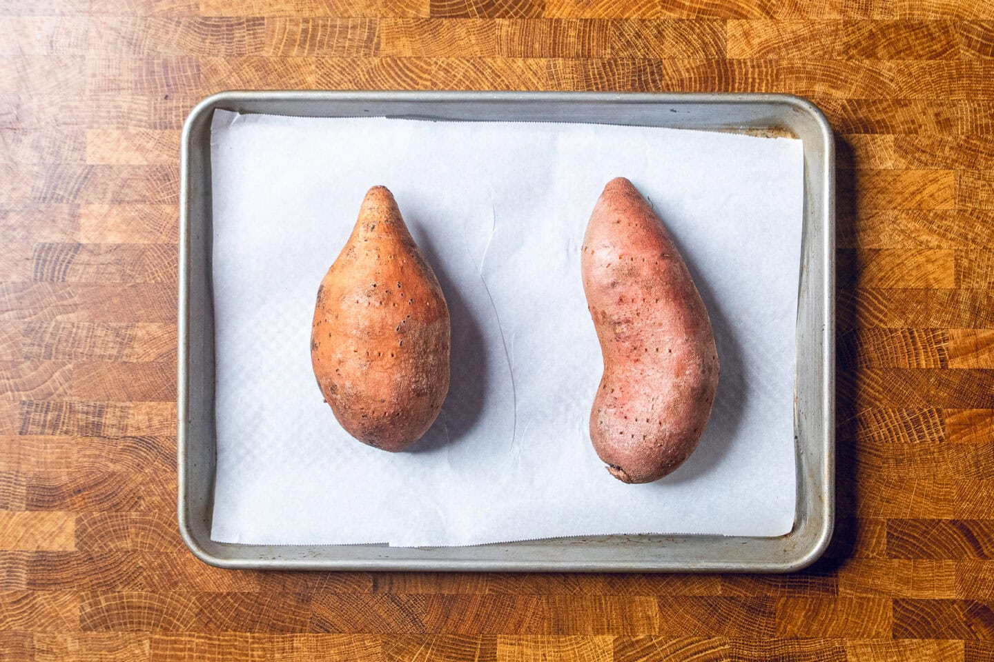 two sweet potatoes with fork holes all over laying on a parchment lined baking sweet before baking.