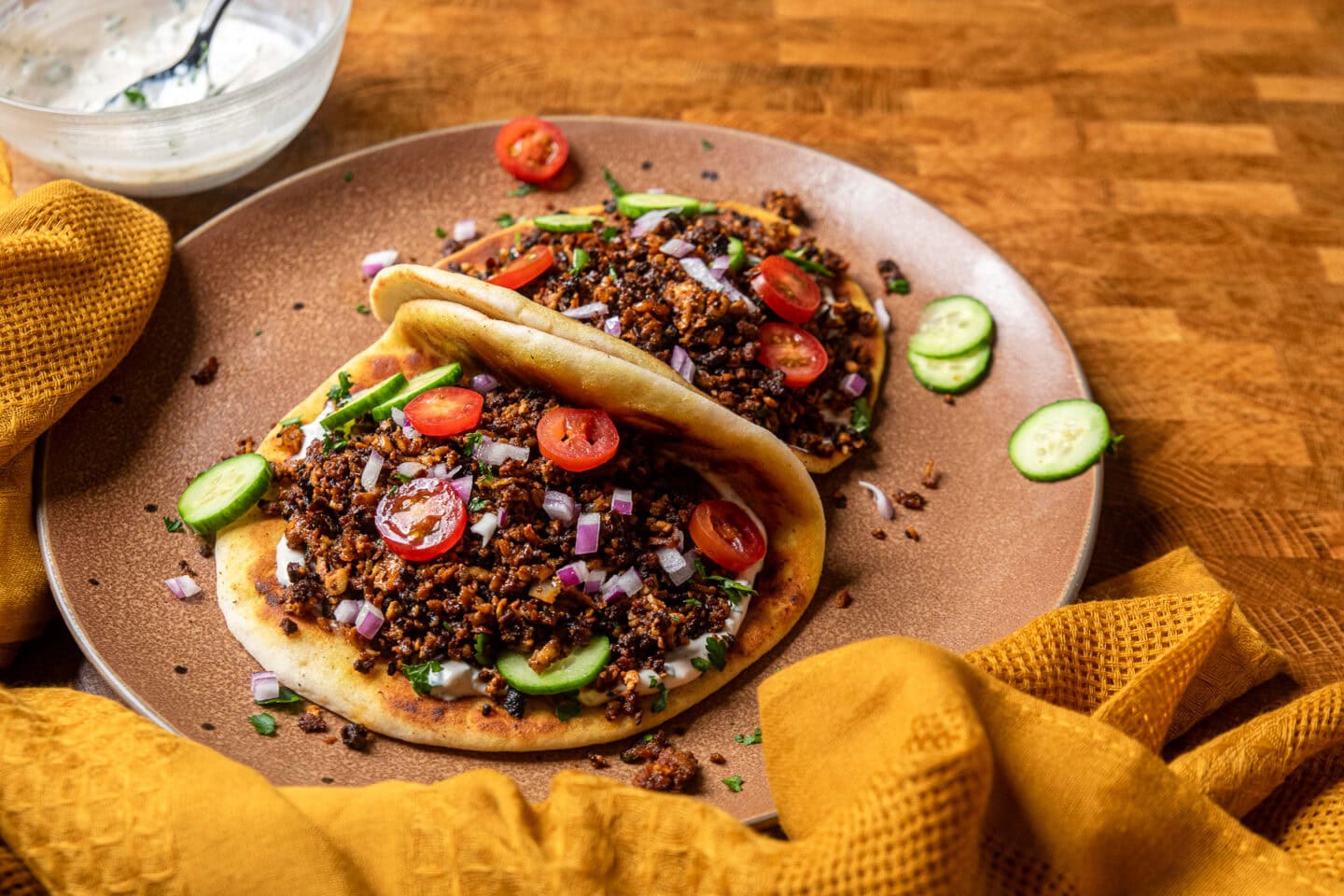 mediterranean baked tofu crumbles on a pita shell with tomatoes, cucumbers, and red onions laying on a brown plate.