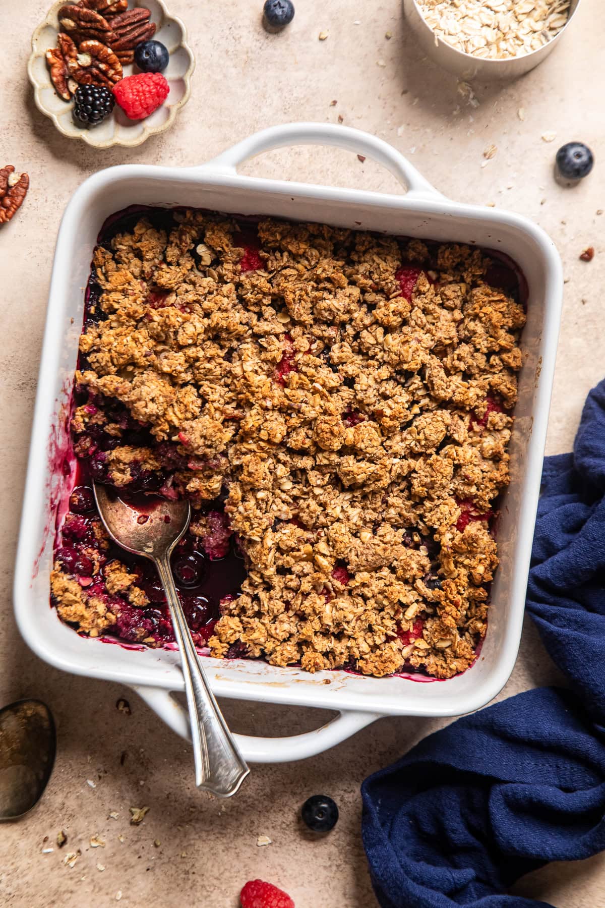 a mixed berry crumble in a white, square baking dish with a piece removed and a spoon resting inside.