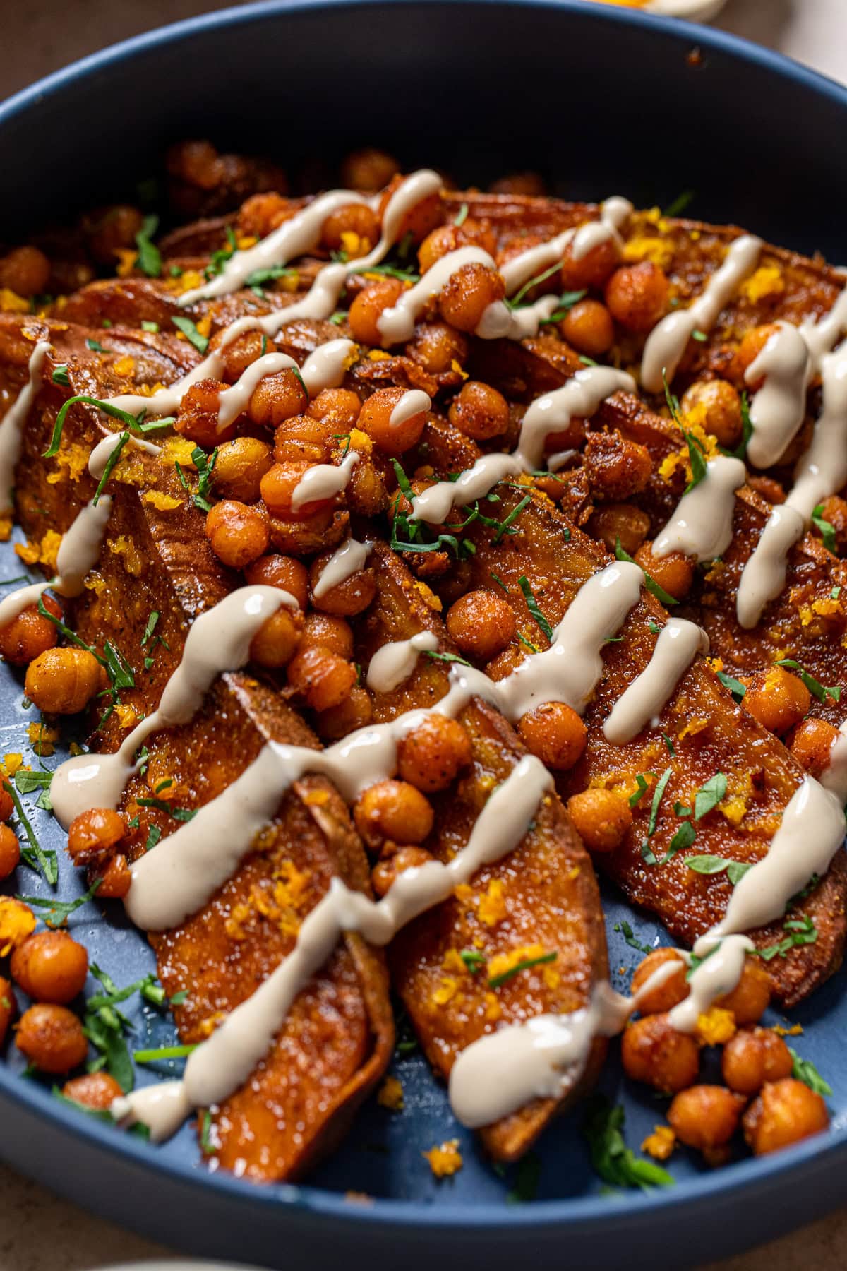 close up of tahini butter sauce over roasted sweet potatoes and crispy chickpeas in a blue bowl.