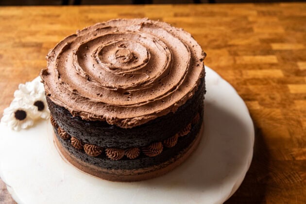 a vegan chocolate cake sitting on a marble lazy susan where the two layers have dollops of chocolate frosting between and a swirled pattern on table, but no frosting on the sides of the cake.