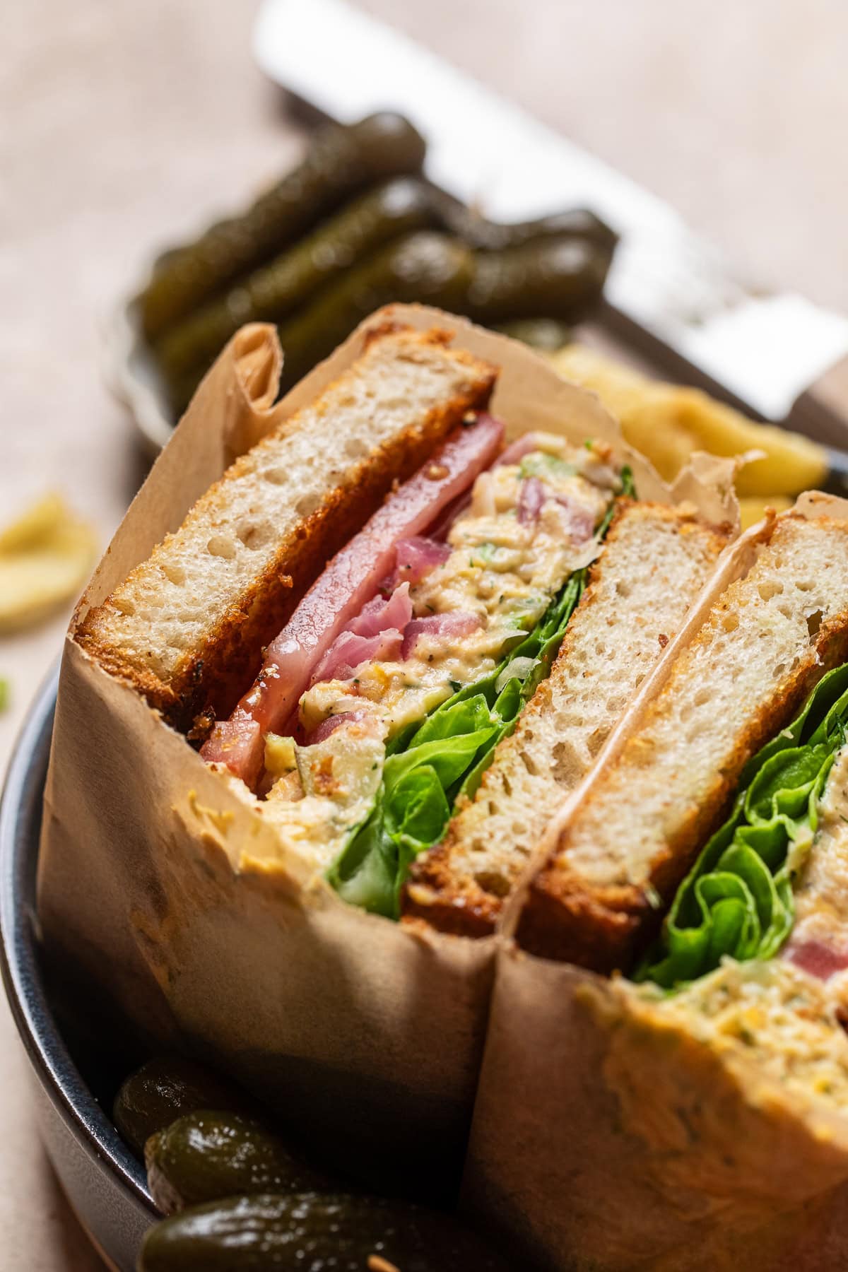 a close up of crispy lemon dill pickle chickpea salad sandwich with lettuce and tomatoes split in half sitting in a bowl.
