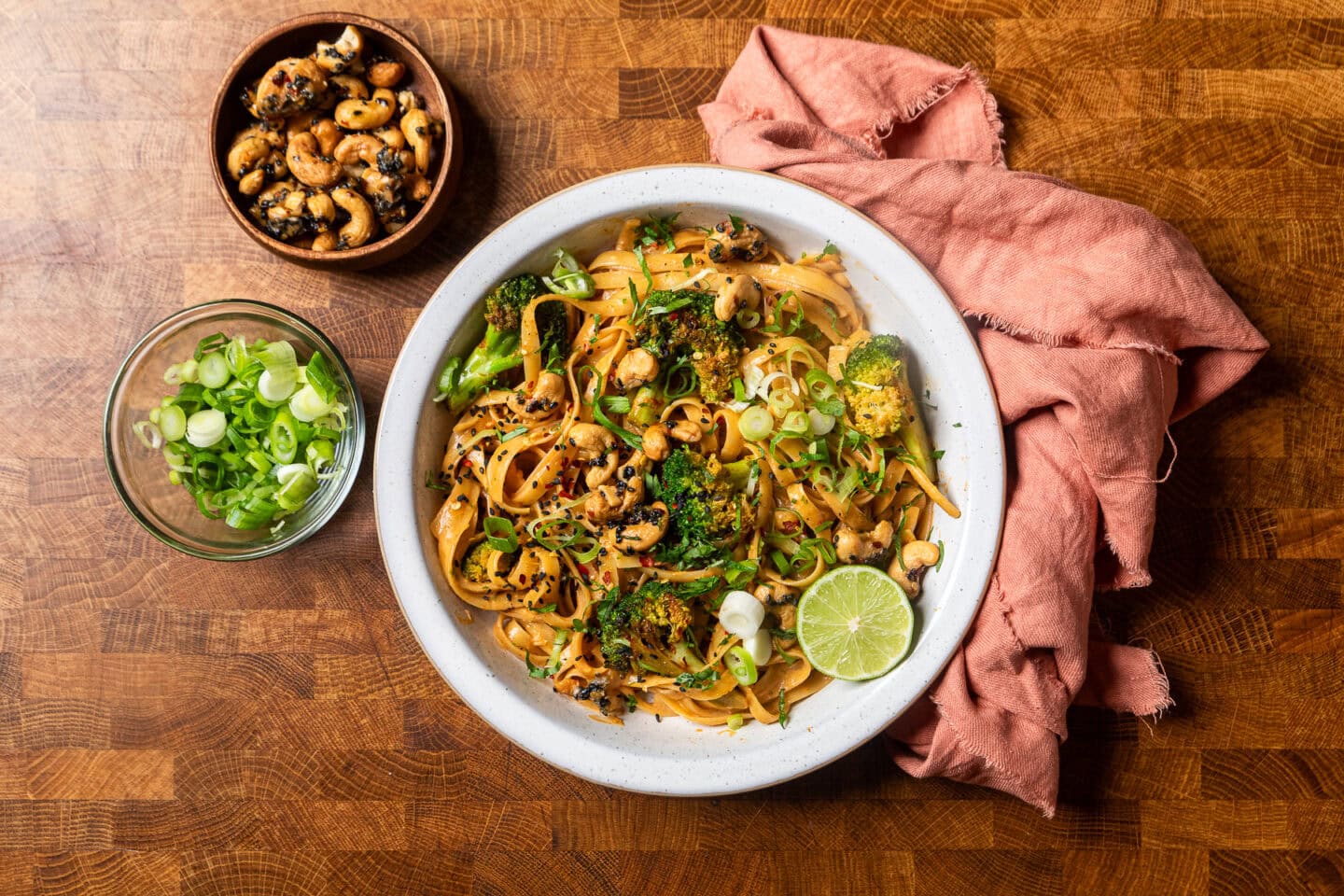 smokey chili lime tahini broccoli noodles in a white bowl with a peach towel on the side and two bowls filled with green onions and candied sesame cashews.