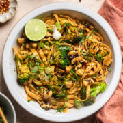 smokey chili lime tahini broccoli noodles in a white bowl with a peach towel on the side and two bowls filled with red pepper flakes and candied sesame cashews.