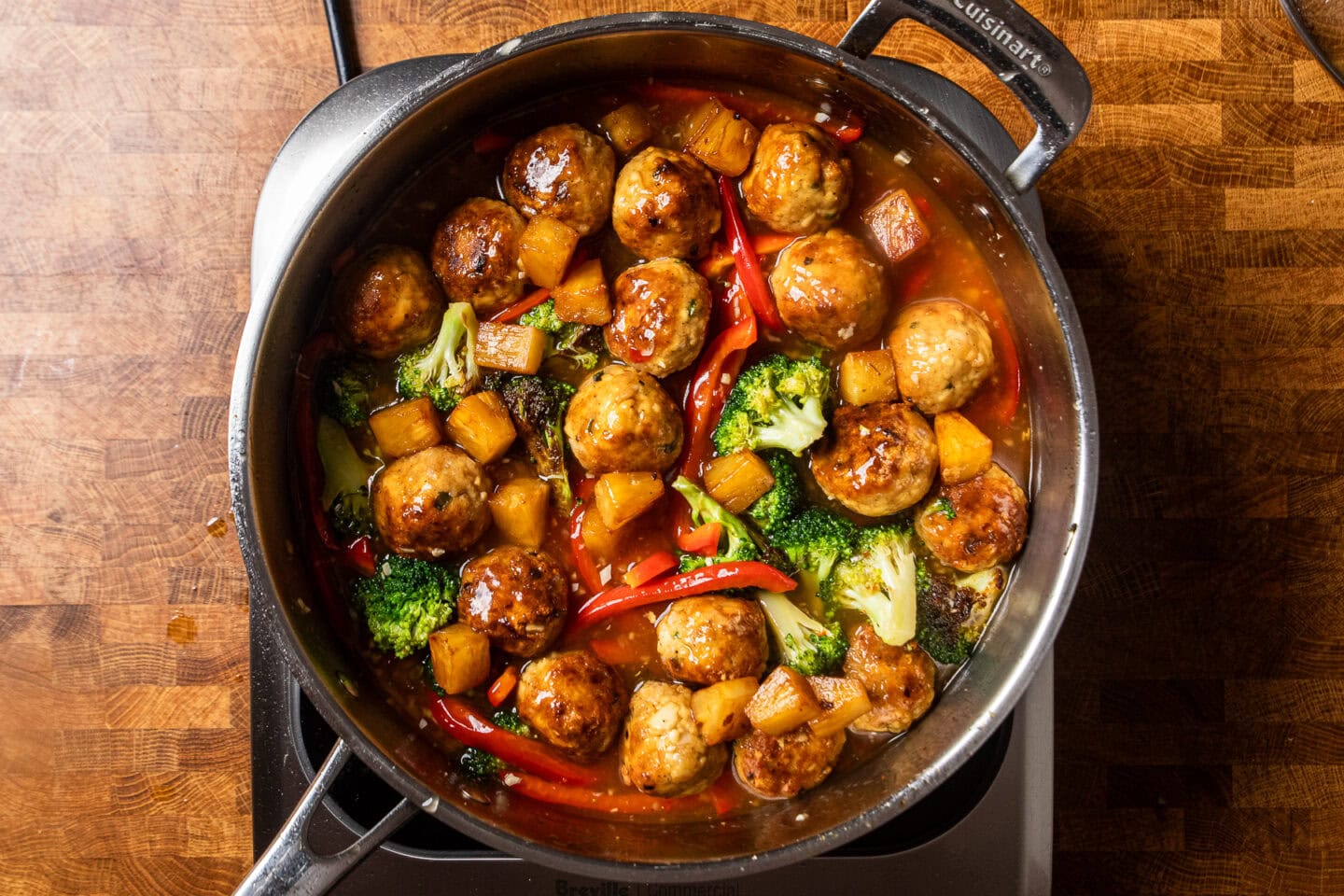 a stainless steel skillet filled with cook tofu meatballs, red pepper strips, pineapple chunks, and broccoli in a sweet chili sauce.