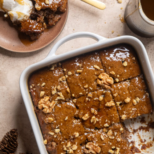 vegan toffee pudding topped with walnuts inside of a square white baking dish next to a plate with some of the pudding and ice cream on it.